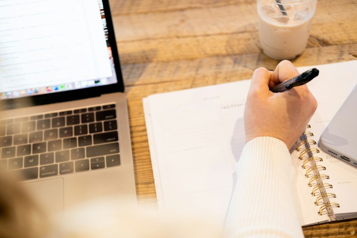 Hånd med penn som skriver i en ringnotatbok ved siden av en oppslått laptop på et trebord